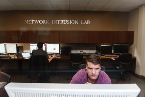 A young man works on a computer. In the background is another young man working on a computer and a sign reading "Network Intrusion Lab".